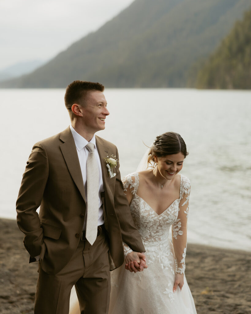 A bride and groom hold hands and laugh in their wedding attire as they walk along the shore at their Lake Crescent wedding