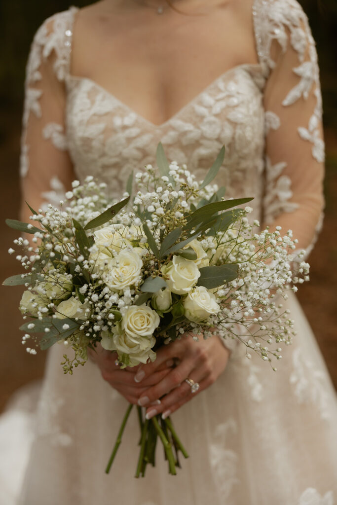 A close-up photo shows a bride in a lace wedding gown holding her bridal bouquet made up of white roses, eucalyptus, and baby's breath at a Lake Crescent wedding