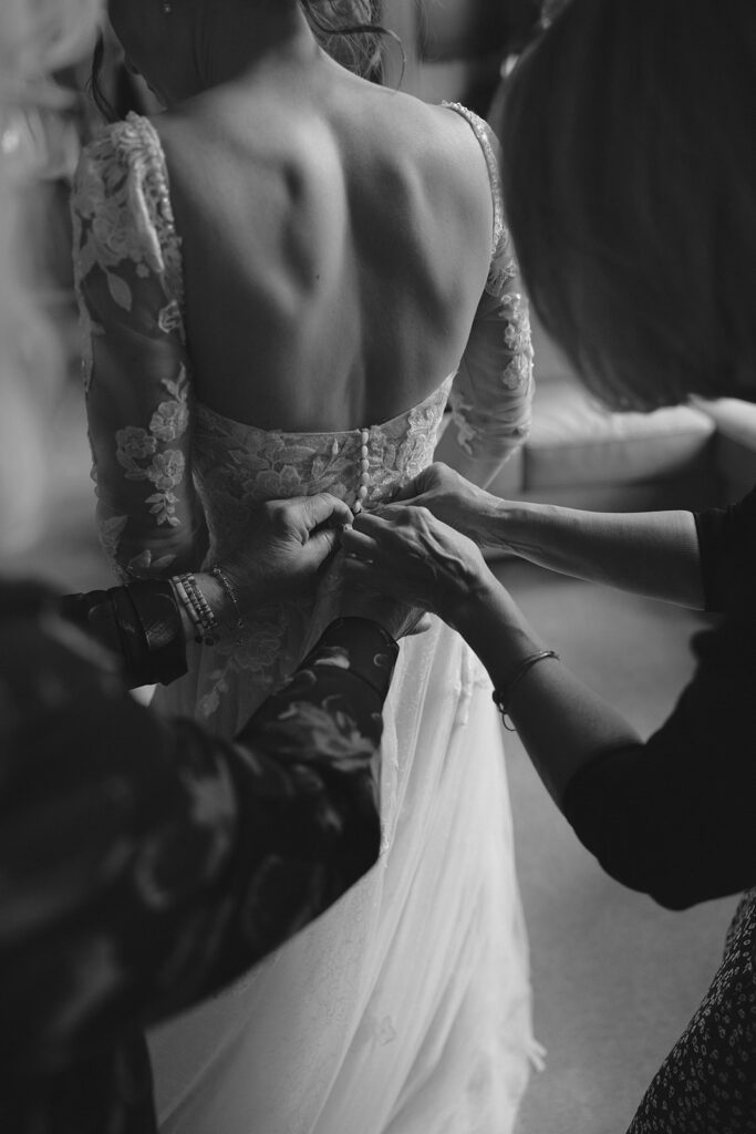 A black and white image of a bride being buttoned into her wedding dress before her Olympic National Park wedding ceremony