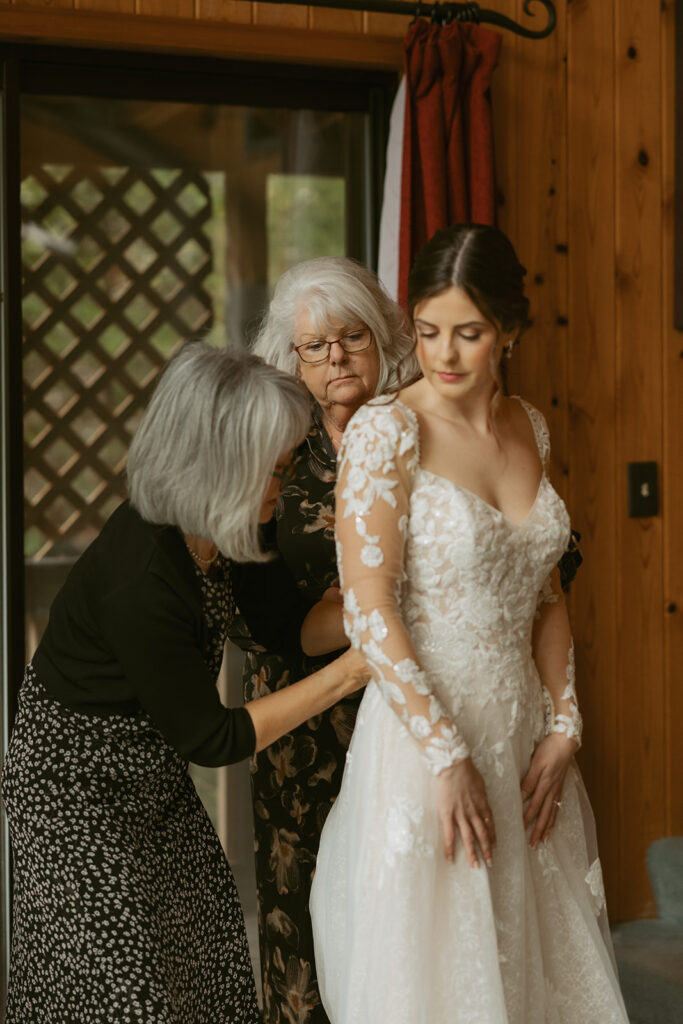 Both of the mother of the bride and the mother of the groom assist a bride with the buttons on her lace wedding gown in a photo taken by olympic national park elopement photographer, Tessa Marie Collective