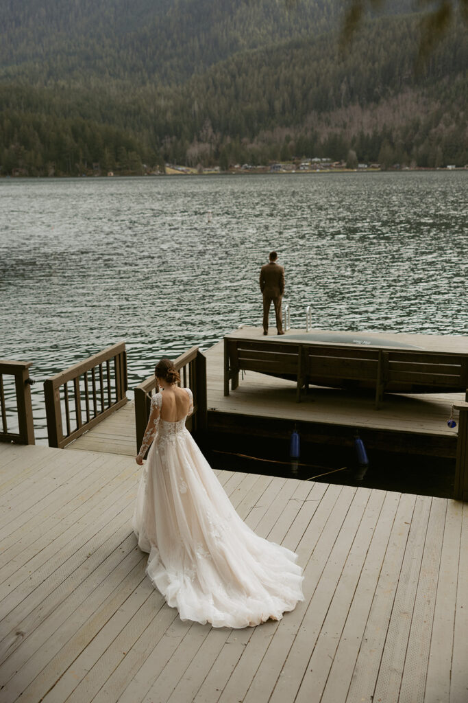 A bride approaches her awaiting groom on the dock of their airbnb for a first look photo before their PNW Elopement ceremony at Lake Crescent