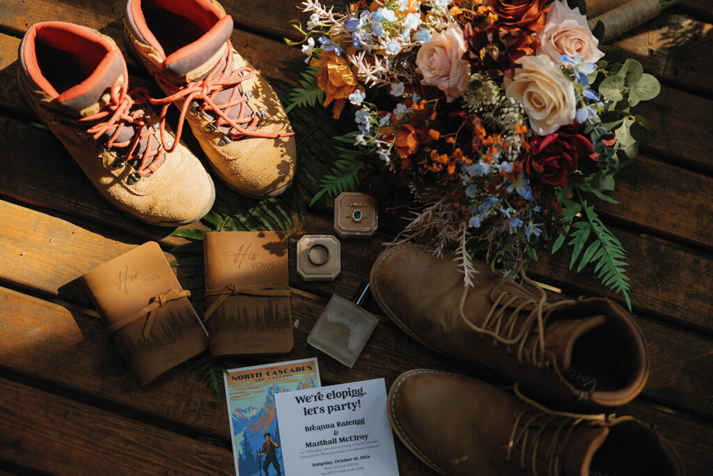 A flat lay of wedding details featuring hiking boots, a bouquet, his & her vow books, wedding rings, and an invitation for a Washington elopement