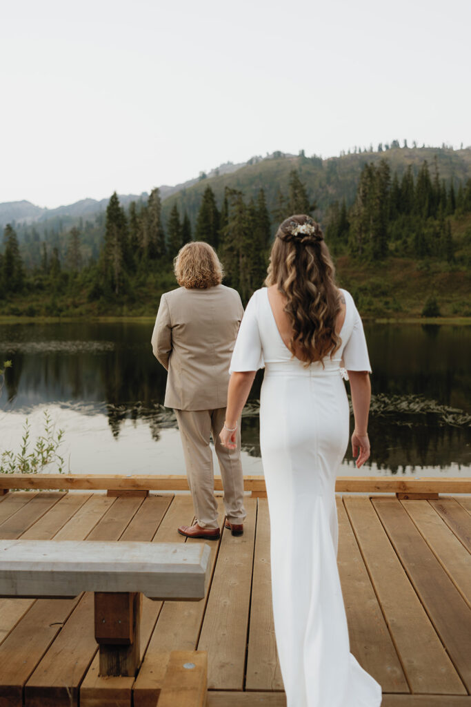 A bride approaches her groom from behind overlooking a scenic lake for a first look before their Washington elopement