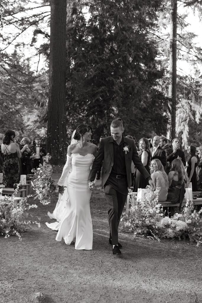 A black and white image shows a bride + groom holding hands as they walk down the aisle at their outdoor wedding in portland oregon