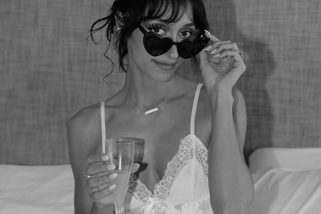 A black and white image shows a bride wearing heart sunglasses holding a mimosa as she gets ready for her Hoyt Arboretum Portland Wedding