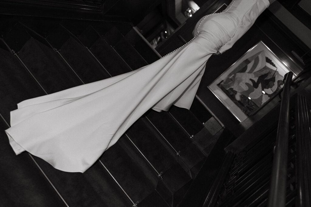 A black and white photo shows a detail shot of a white wedding gown with buttons down the back as the bride wearing it stands on a staircase at The Sentinel Portland