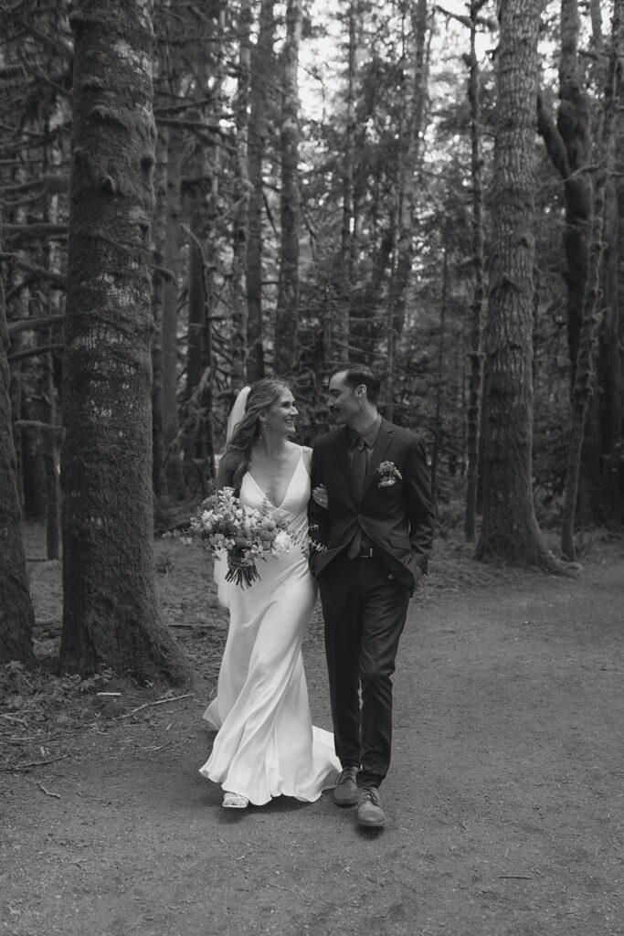 A black and white photo shows newlyweds smiling at each other as they walk through the forest at their national park elopement at Lake Crescent