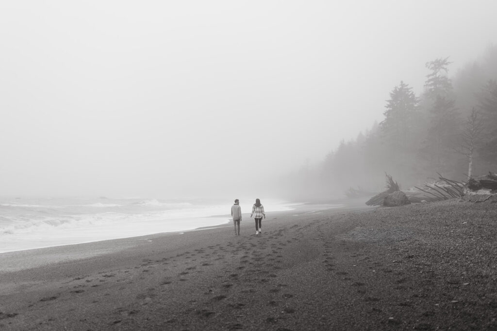 A black and white image shows a couple walking along the shore at Rialto Beach during a photoshoot to prep for their Olympic National Park Wedding
