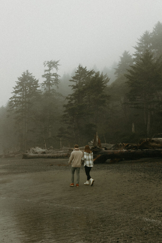 A couple walks hand in hand away from the camera in a photo taken on a misty day at Rialto Beach in preparation for their Olympic National Park wedding