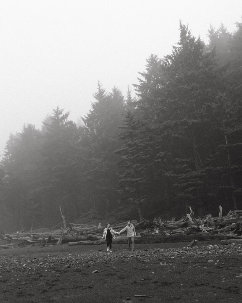 A black and white image of a couple walking hand in hand along the shore at Rialto Beach in a photo taken by an Olympic National Park Elopement Photographer