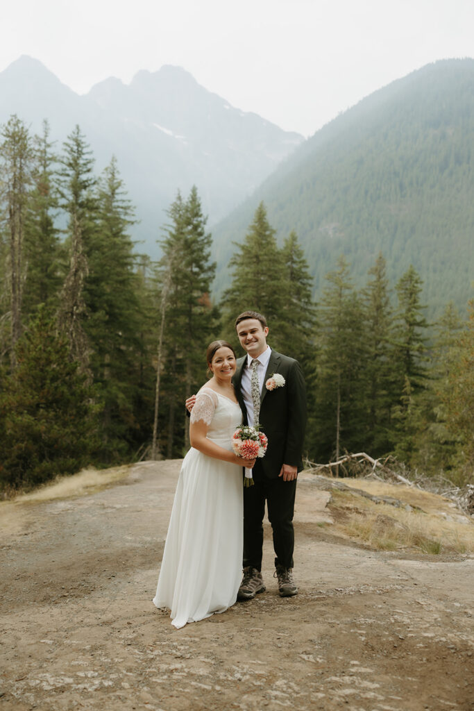 A groom wearing a suit and hiking boots stands with his arm around his bride at their WA state elopement