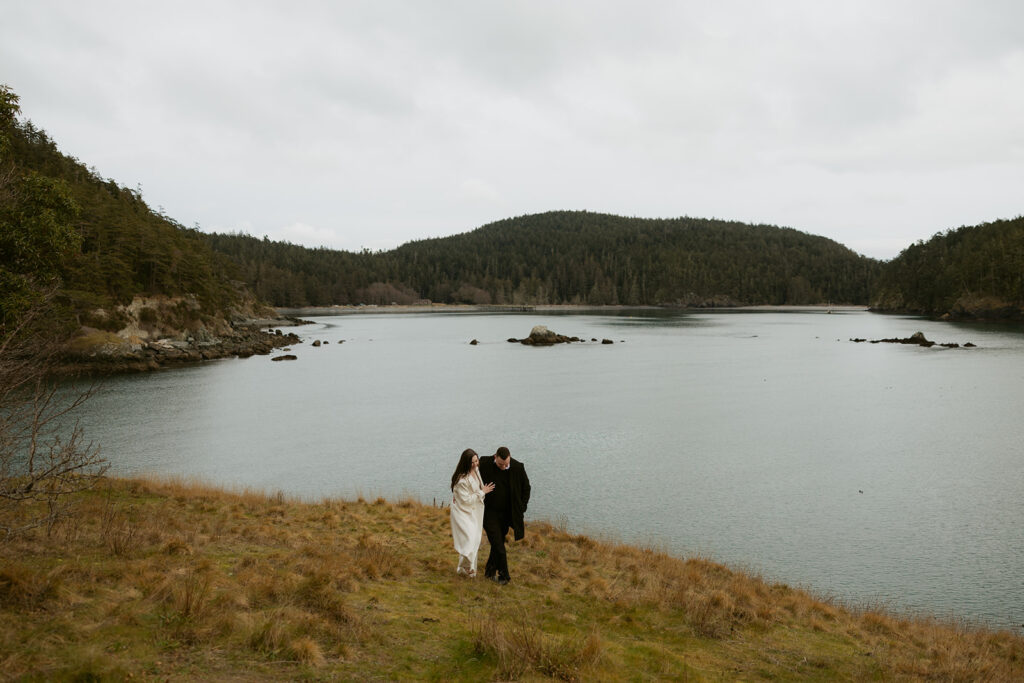 A newly married couple embrace as they walk along the waters edge at Rosario Lake in a photo taken by Washington elopement photographer, Tessa Marie Collective