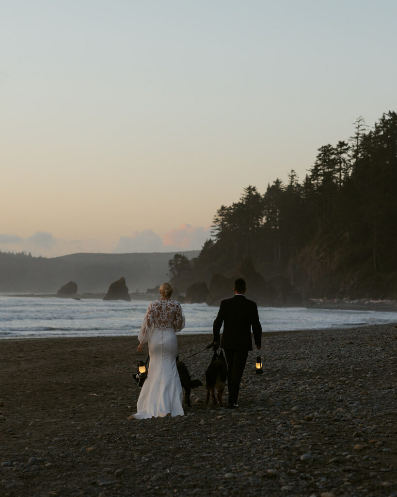 A bride and groom walk along the shore with lanterns in hand at dusk following their Ruby Beach Washington elopement