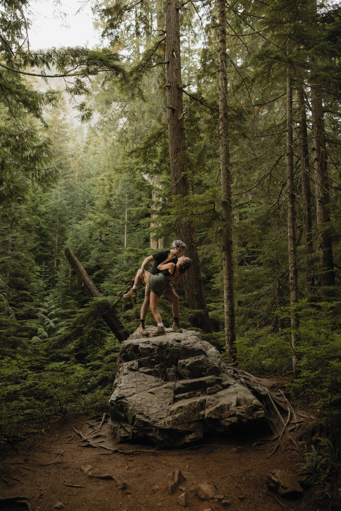 A couple share a kiss at op a boulder on a hike during their Washington engagement photos 