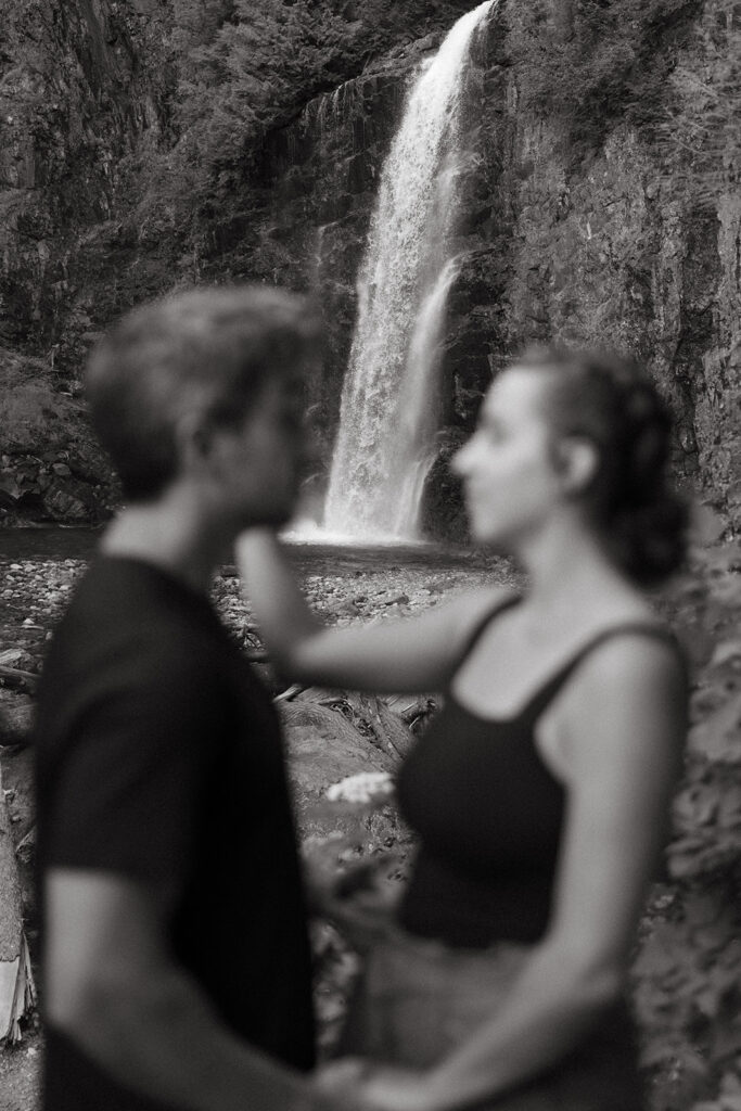 A black and white image shows an out of focus couple standing before the focal point of Franklin Falls WA 