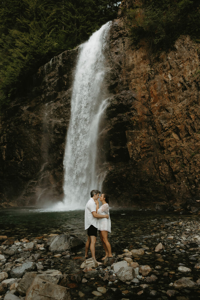 A couple wearing white embrace in the water beneath Franklin Falls during their Washington engagement photos 