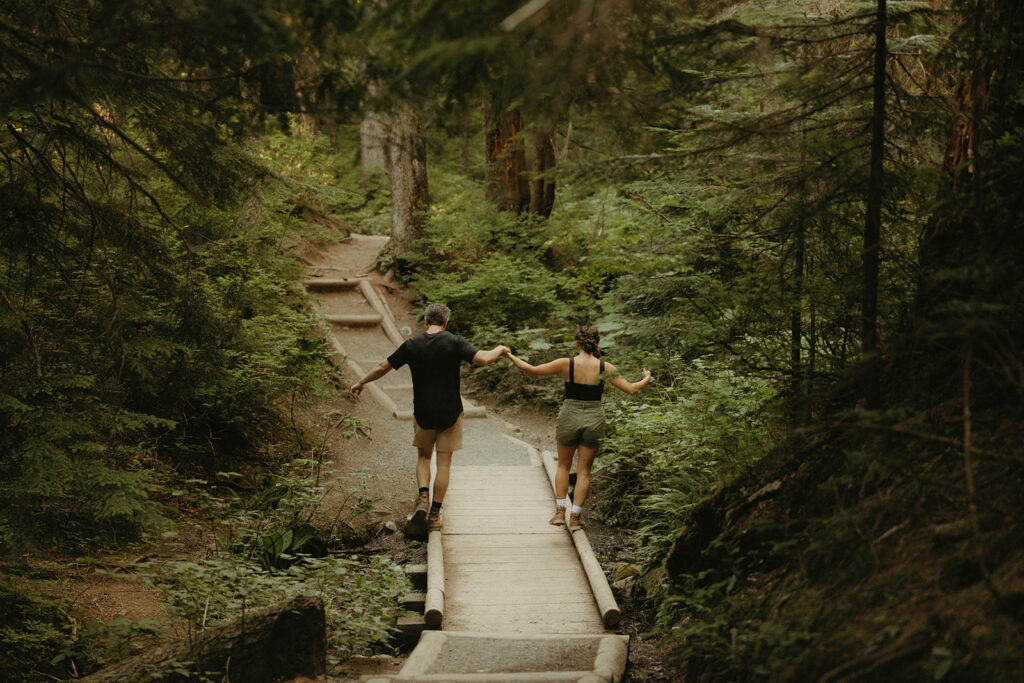 An engaged couple hold hands as they balance on the edges of a wooden bridge during Washington engagement photos taken at Franklin Falls 