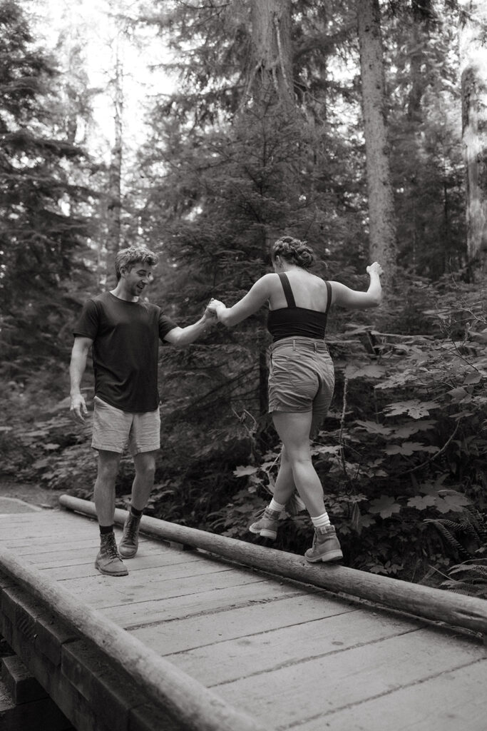 A black and white image shows a man holding his fiancée's hand as she balances on a bridge railing in Seattle engagement photography
