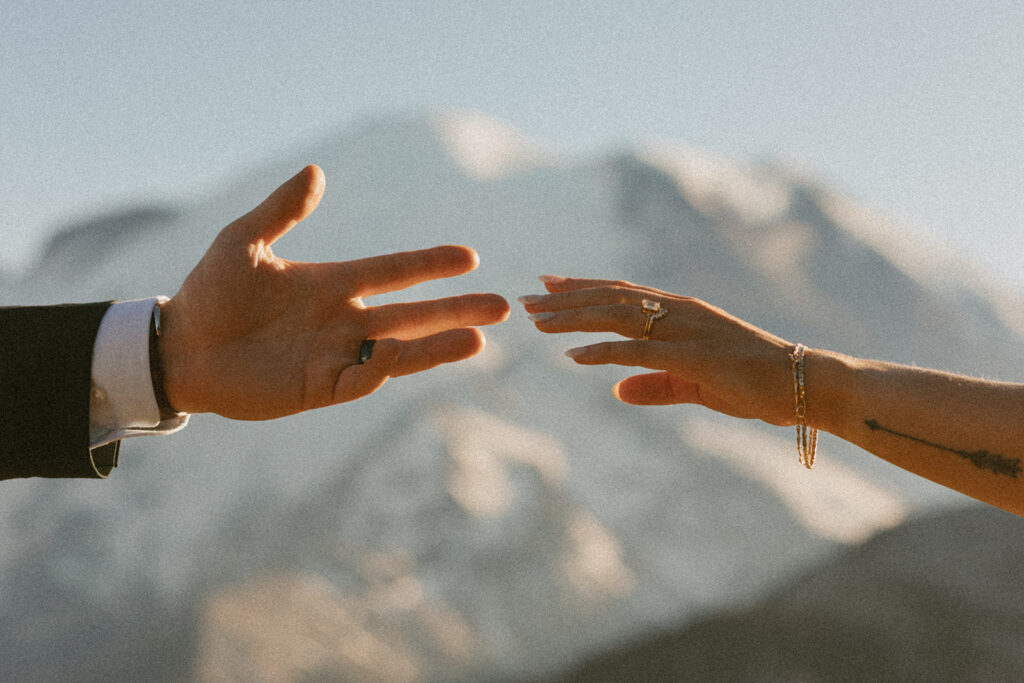 A newly married couple reach for reach other, showing off their wedding bands with mountains in the background at their Mount Rainier elopement 