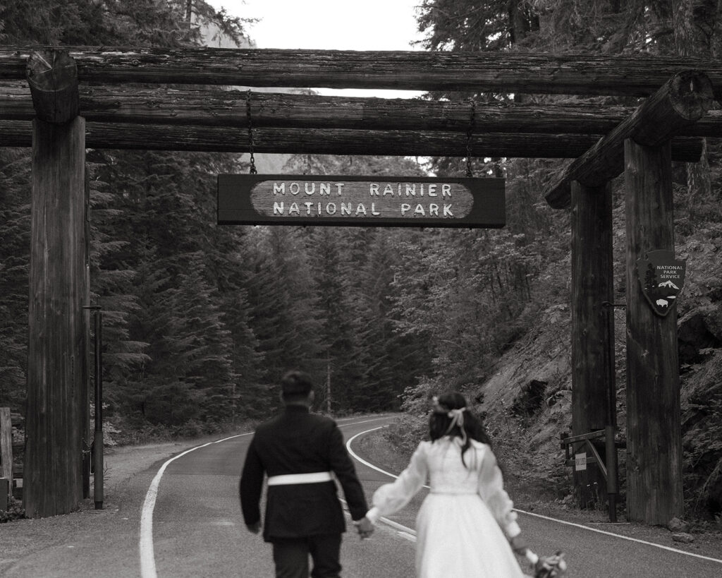 Newlyweds hold hands as they walk underneath a sign in a b+w photo taken at their mount rainier elopement