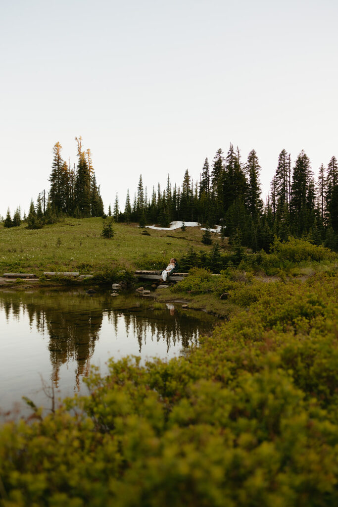 Newlyweds are photographed at a distance seated by a lake surrounded by alpines in an image taken by their mt rainier wedding photographer 
