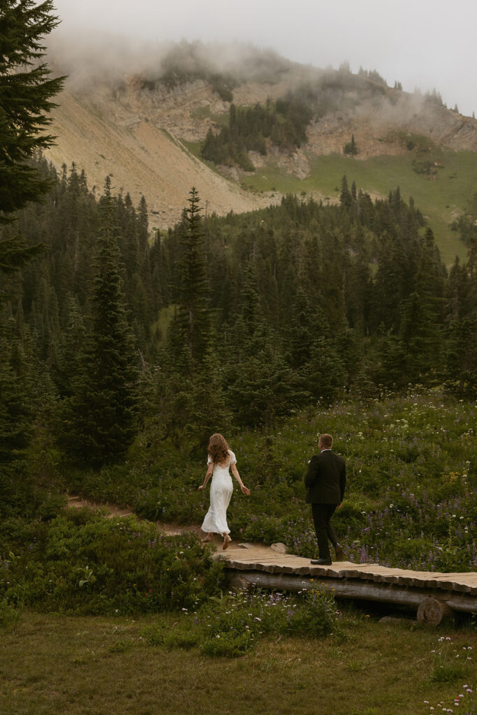 A bride and groom in their wedding attire walk along a wooden bridge in the forest at their Mt. Rainier wedding 