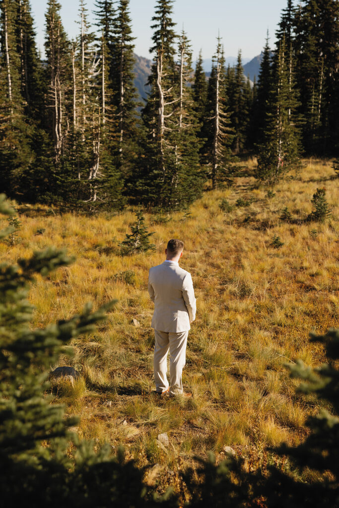 A groom in a tan suit is shown standing amongst the alpines at his mount rainier elopement