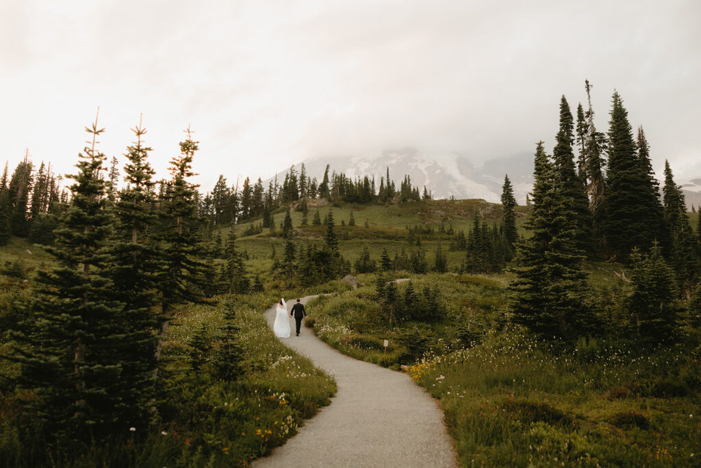 A bride in her gown and a groom his suit hold hands as they walk along a trail following their Mt Rainier elopement 