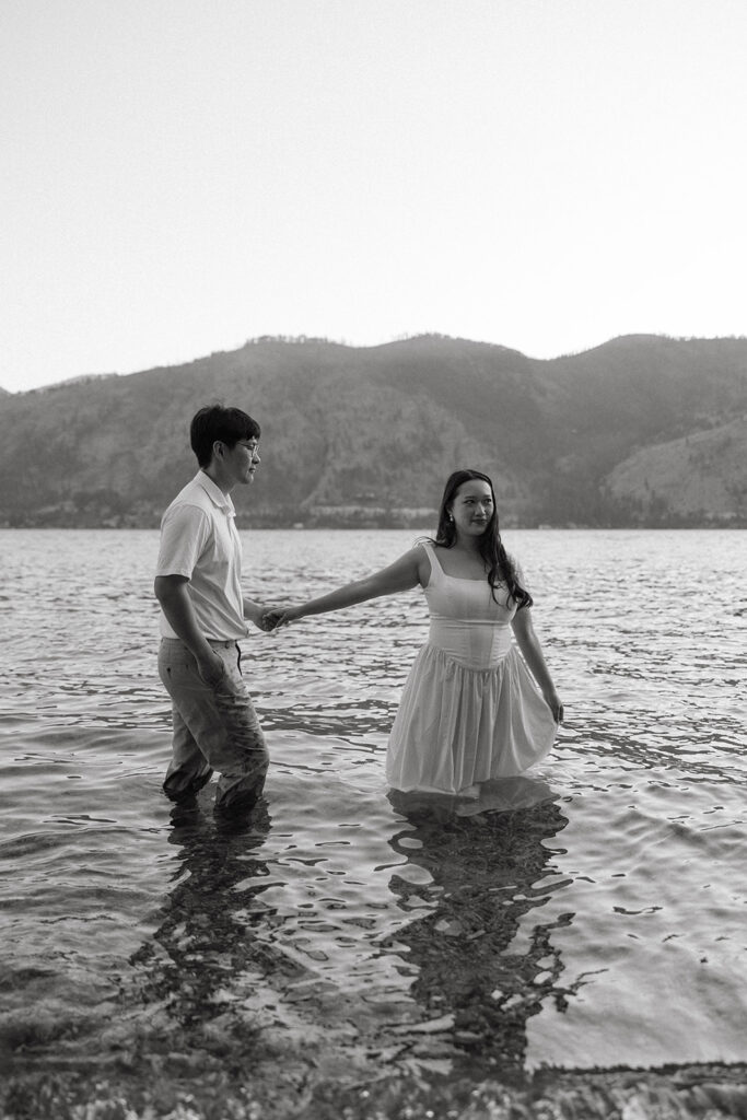 A b+w image shows a couple wading in the water at Lake Chelan during their Willow Point Park engagement session in Manson, WA