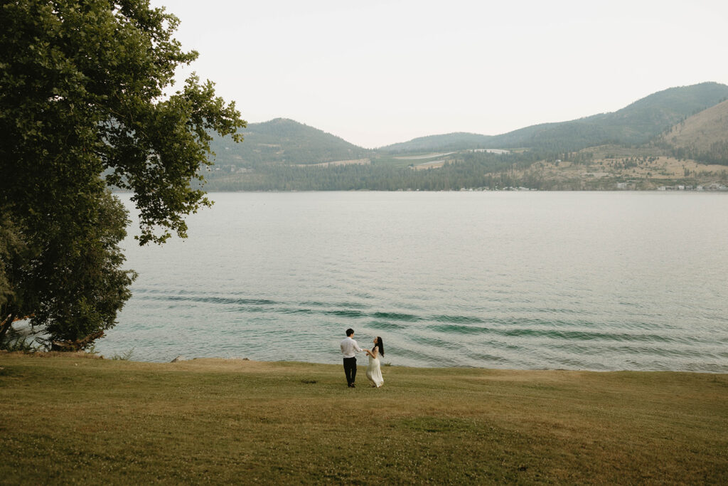 A newly engaged couple is photographed from a distance with Lake Chelan in the background during a willow point park engagement session