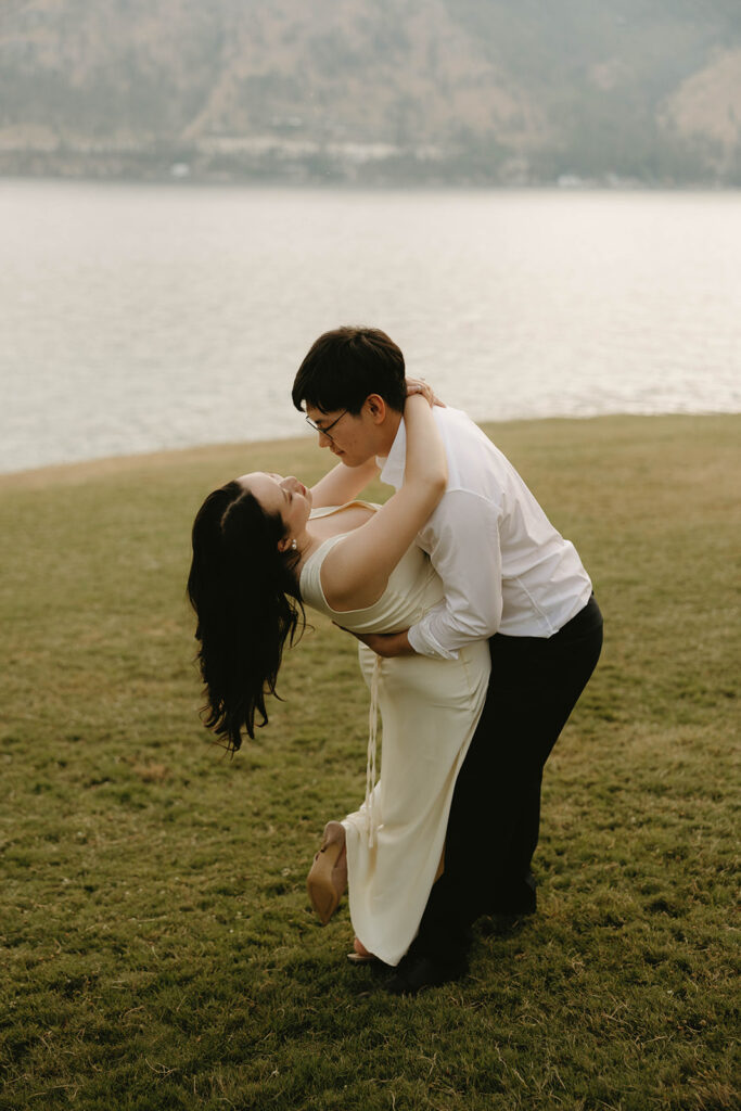 A man wearing a white shirt and black pants dips his fiancee in a lakeside lake chelan engagement session