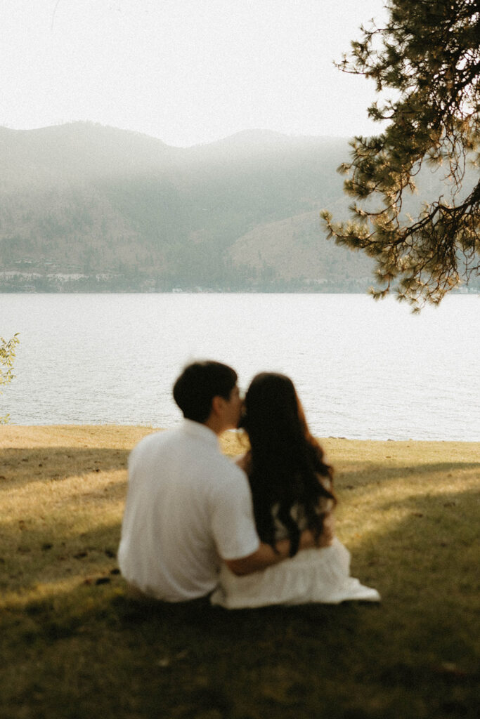 An out of focus couple share a kiss with Lake Chelan in the background during engagement photos taken in Manson Washington