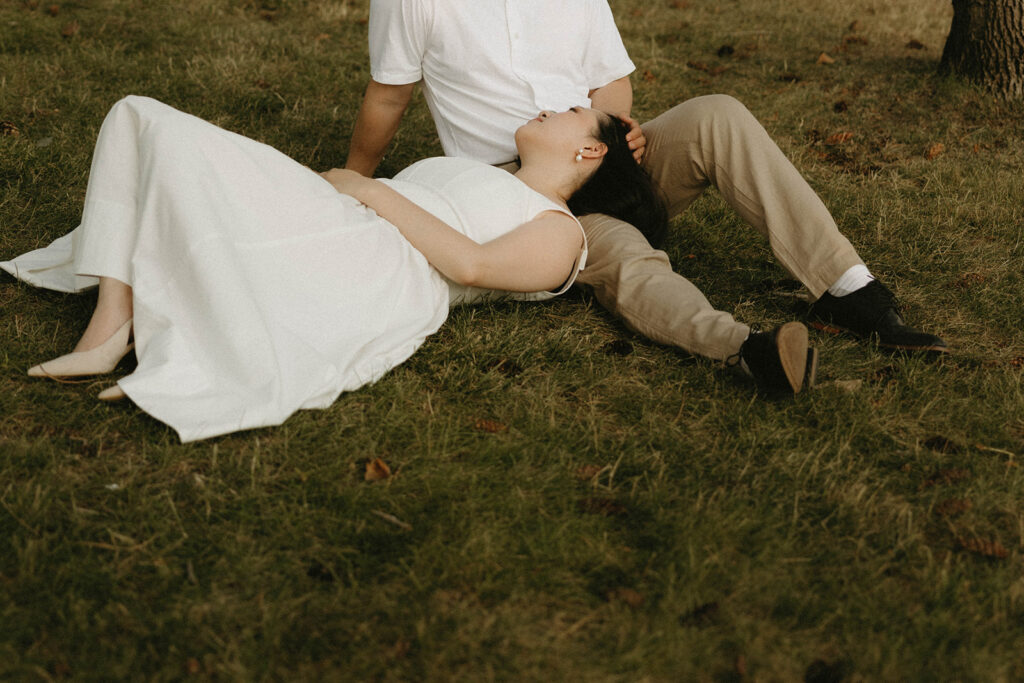 A woman wearing a white dress rests her head on her fiance's lap during a lakeside lake chelan engagement shoot