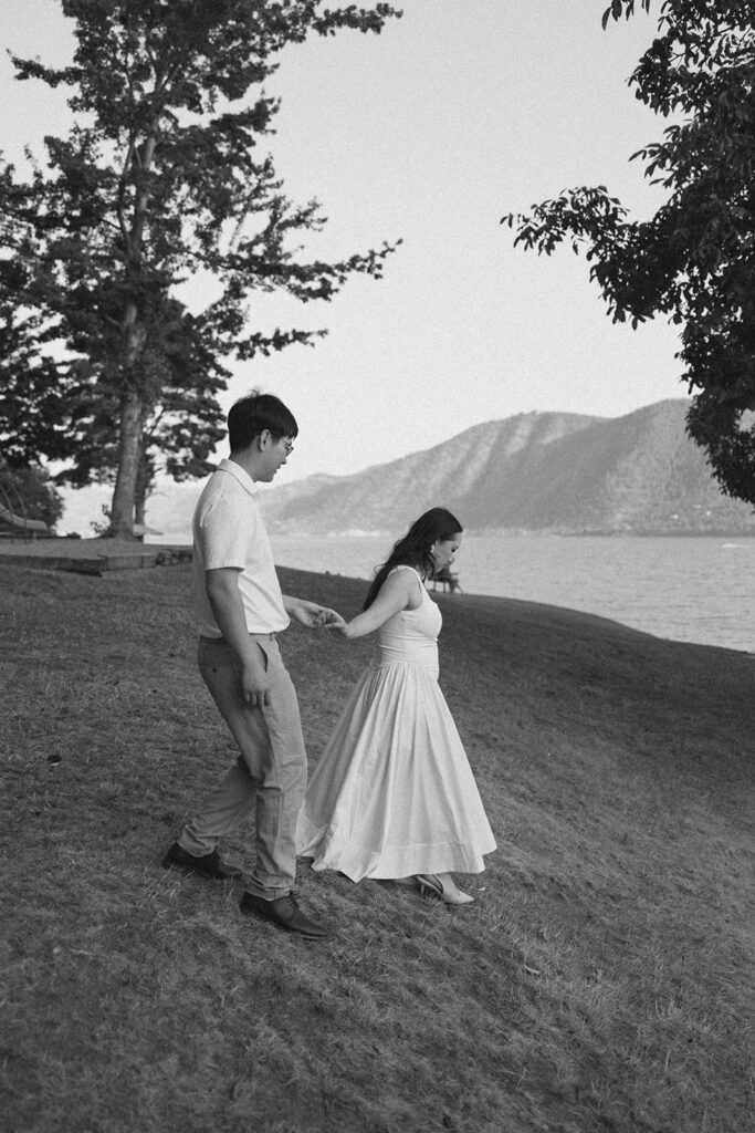 A b+w image shows an engaged couple holding hands as they walk toward the water at Willow Point Park in Manson WA