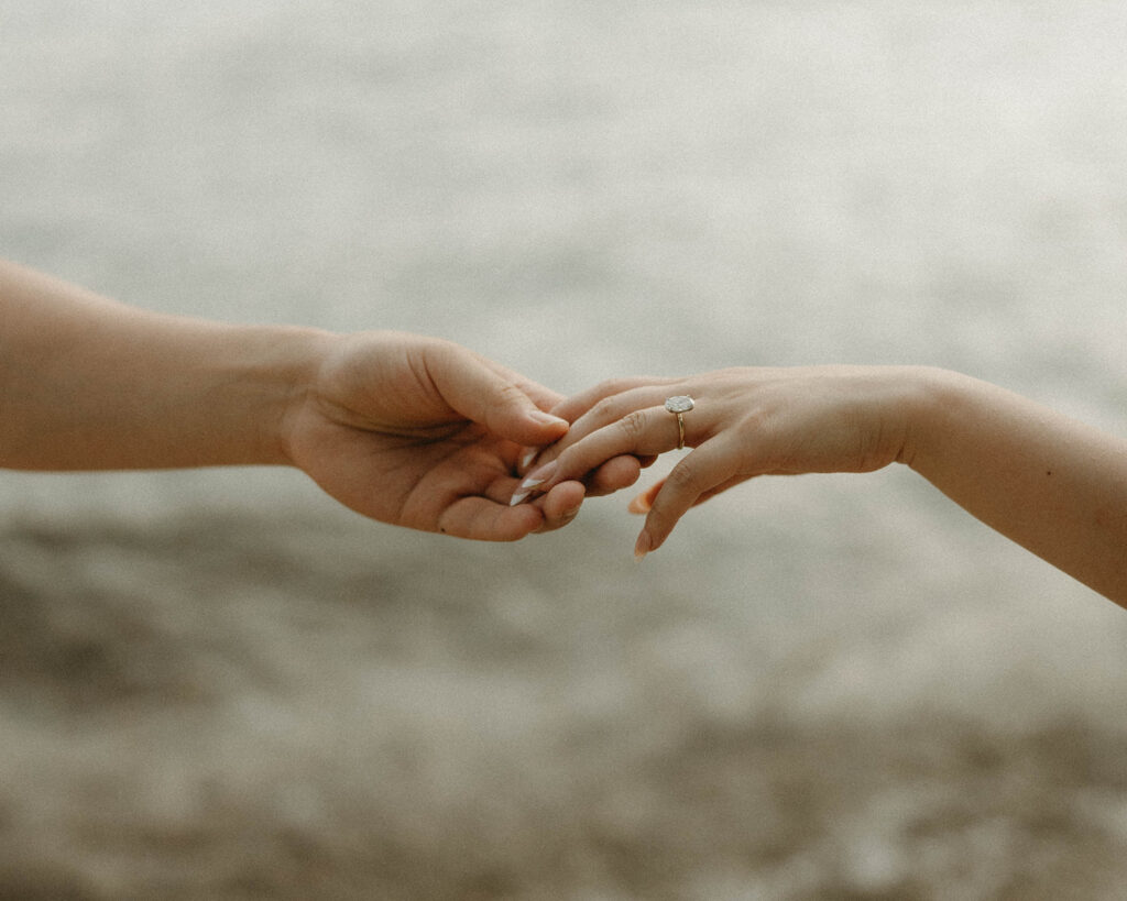 A close up photo shows a couple holding hands with a focus on a diamond engagement ring on a thin gold band during an engagement photoshoot with a lake chelan photographer