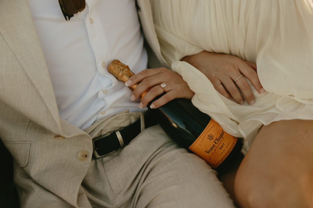 A newly engaged couple holding a bottle of champagne showing off the engagement ring